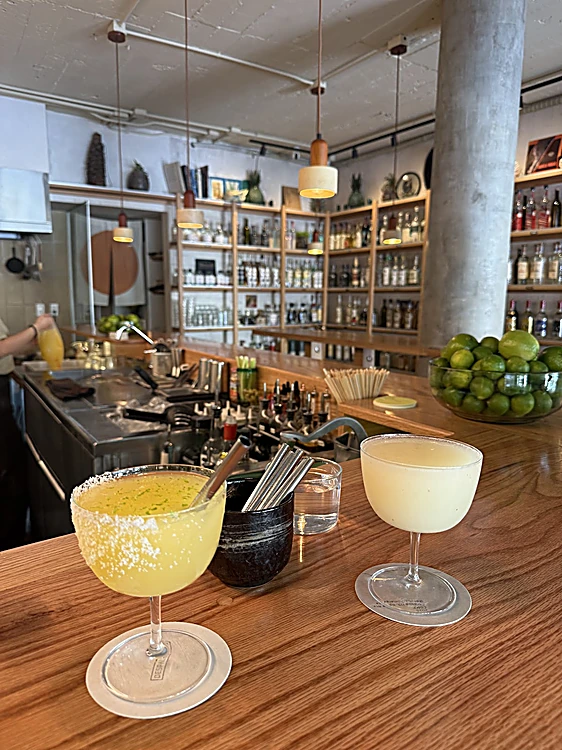 Dos margaritas en copa sobre la barra de roble, con limones y los estantes al fondo, de día