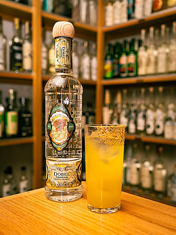 Botella de tequila y un trago amarillo con escarcha frente a los estantes de roble llenos de botellas