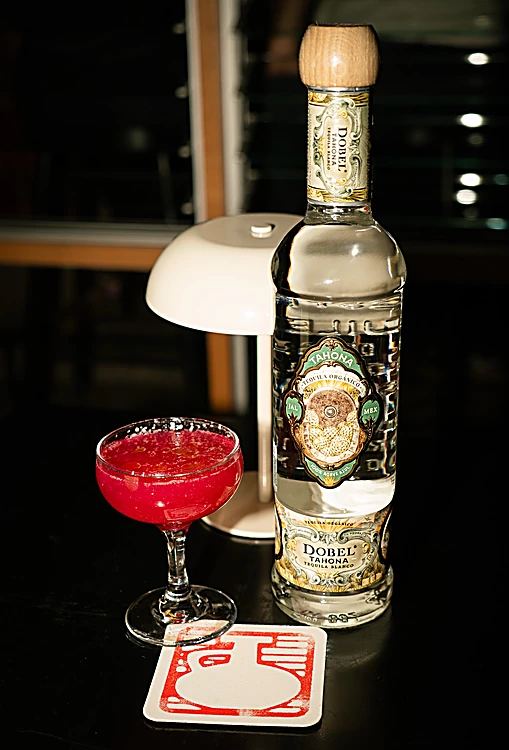 Margarita rosa en copa junto a una botella de tequila y un portavasos rojo, de noche