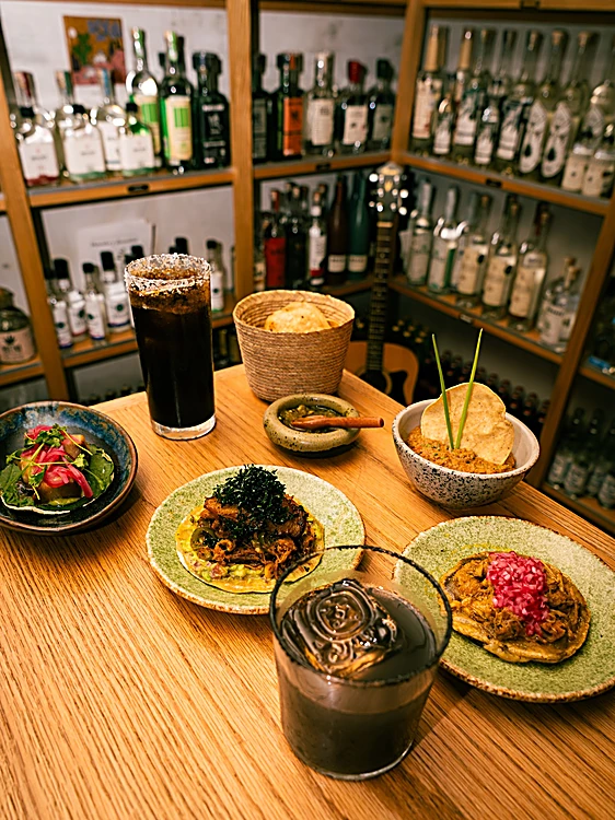 Barra con varios platos para compartir y tragos frente al estante de botellas
