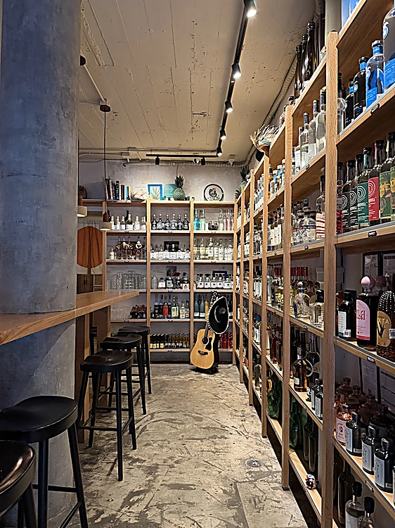 La sala de Despacho de día: estantes de roble de piso a techo llenos de botellas, una guitarra en el estante y bancos altos junto a la barra de pared